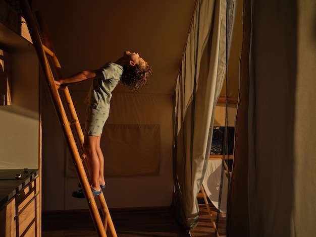 Side view of child in pajama holding onto ladder and looking up in kitchen of cozy glamping tent with wooden furniture during summer holidays