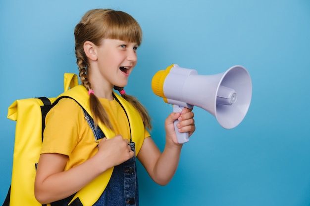 Side view close up of pretty little blonde schoolgirl scream in megaphone, wears yellow backpack, posing isolated on pastel blue background in studio. Childhood lifestyle concept. Mock up copy space