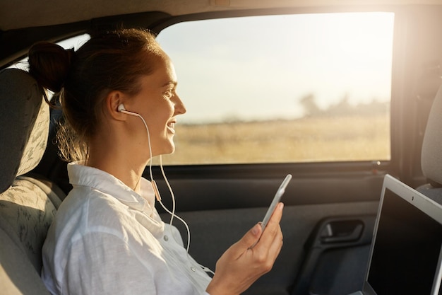 Side view closeup portrait of businesswoman sitting on back seat of car and working with laptop listening music in earphones holding mobile phone in hand looking at window enjoying sunset