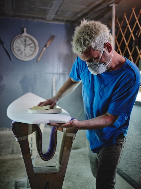 Side view of concentrated blond haired male master in respirator mask polishing white surfboard placed on shaping racks in workshop