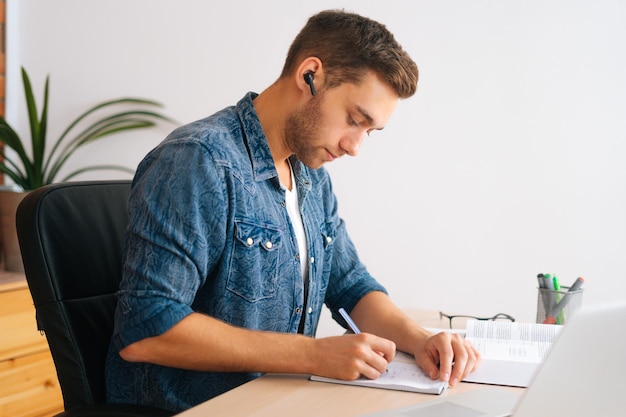 Side view of concentrated male student studying online from home using laptop writing notes, watching video class lesson, listening audio course, studying with teacher distant sitting at desk.