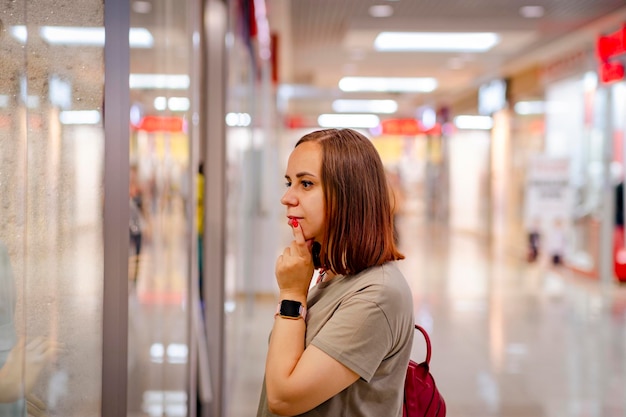 Side view of concentrated young female customer with brown hair in casual clothes and smartwatch standing near showcase during shopping in modern mall