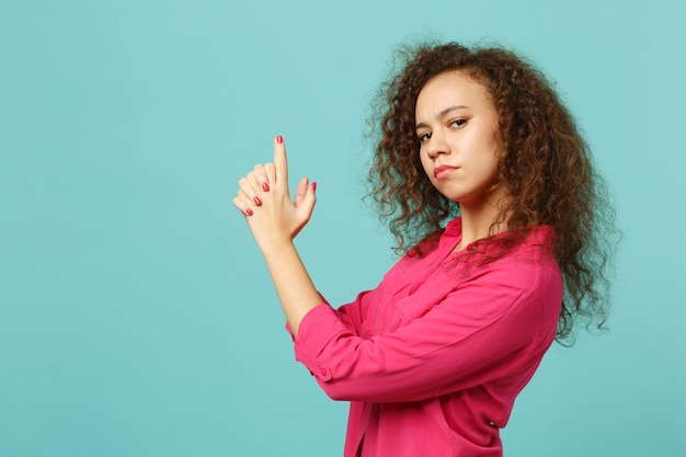 Side view of confident african girl in pink casual clothes holding hands like gun isolated on blue turquoise wall background in studio. People sincere emotions, lifestyle concept. Mock up copy space.