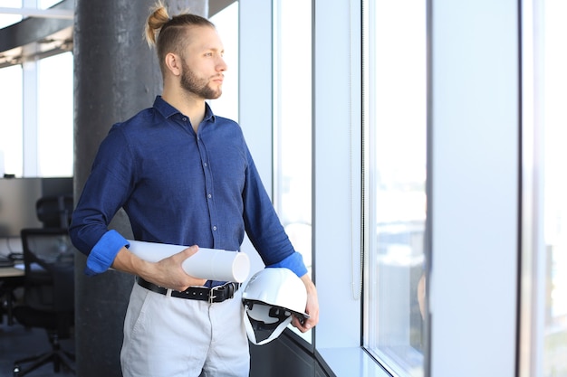 Side view of confident young business man holding blueprint and hardhat and looking away while standing indoors.