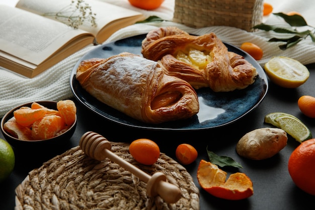 Side view of croissants in plate with tangerine slices and open book with basket of kumquat and little flower on book with ginger lime slices trivet and honey dipper on cloth on black background