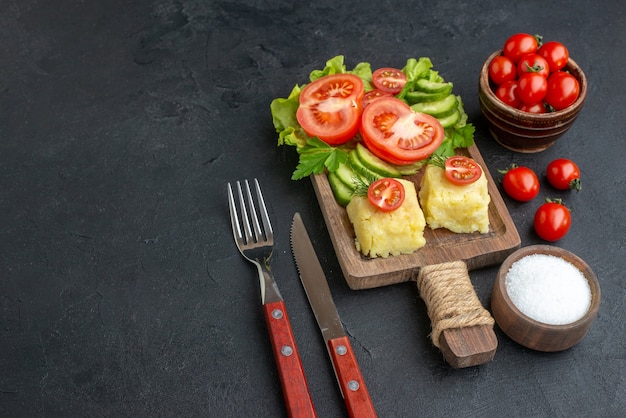 Side view of cut whole fresh tomatoes and cucumbers cheese on wooden board cutlery set salt on black surface