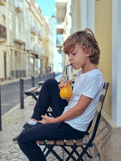 Side view of cute boy drinking fresh juice from bulb shaped bottle while sitting on street near building in city