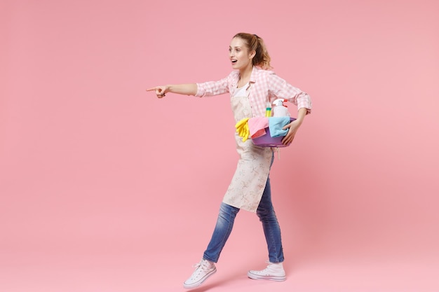 Side view of excited young woman housewife in apron hold basin with detergent bottles washing cleansers doing housework isolated on pink background. Housekeeping concept. Pointing index finger aside.