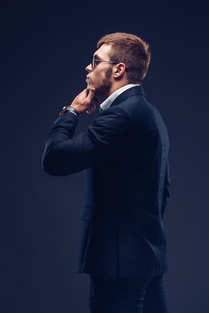 Side view of a fashion bearded young serious man in sunglasses luxury suit with one hand in pocket other is on the chin