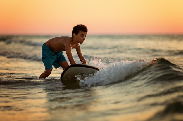 Side view of fearless kid floating surfboard at ocean with waves on sunny evening