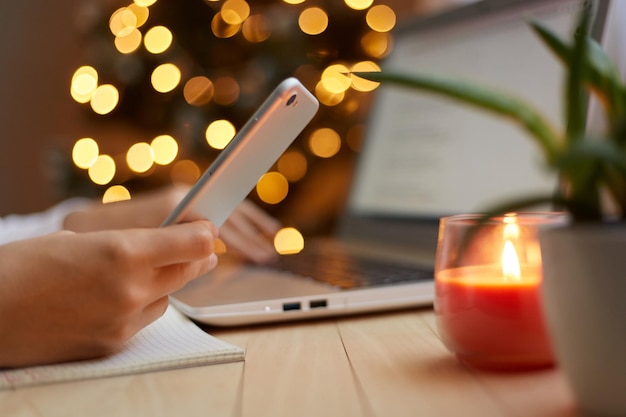 Side view of female person's hand holding mobile phone in hands unknown woman using cell phone browsing internet with blurred Christmas tree on background