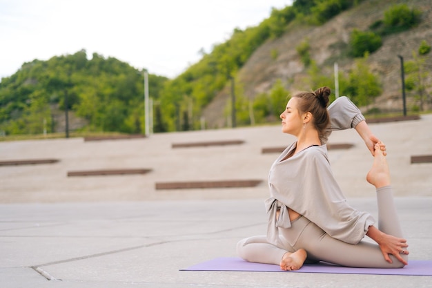 Side view of flexible Caucasian young yogini woman in sportswear performing yoga Pigeon pose outdoors sitting on yoga mat