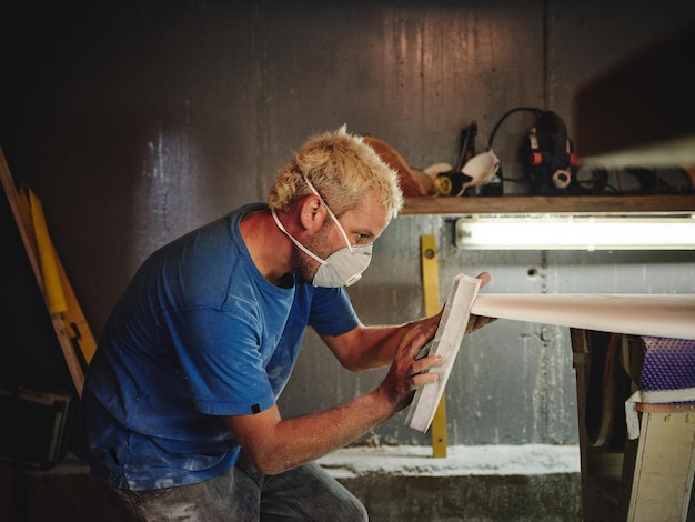 Side view of focused carpenter man in protective respiratory mask polishing nose of surfboard with abrasive tool in workshop