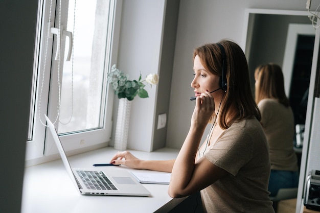 Side view of focused female operator talking using headset and consulting client sitting at window sill with laptop from home office