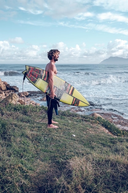 Side view full body of bearded muscular male surfer standing on rocky hill with surfboard in hand and admiring powerful wavy ocean in daylight