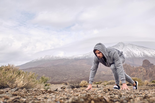 Side view full body of focused Hispanic male traveler in sportswear standing in crouch start position on stony ground against mountain Teide in Tenerife Canary Islands Spain