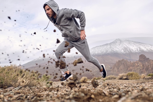 Side view full length ground level of focused Hispanic male athlete in sportswear running fast against mountain Teide with and throwing stones out of way in Tenerife Canary Islands Spain