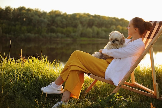 Side view full length portrait of happy young adult woman wearing white shirt and her Pekingese dog sitting at sunset near the river enjoying beautiful view