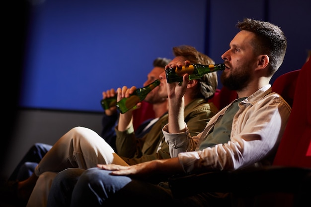 Side view at group of men watching movie in cinema and drinking beer while sitting in row on red seats, copy space