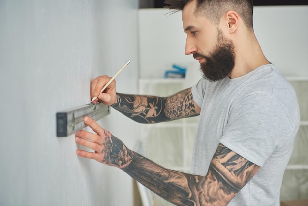 Side view of handsome bearded young man holding level tool and pencil during home improvement