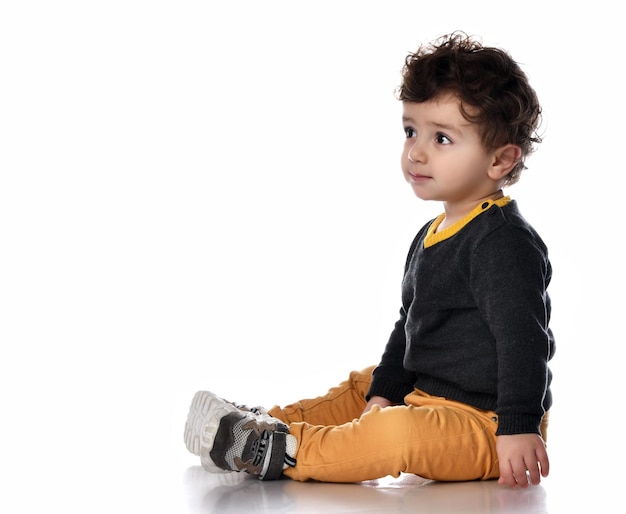 Side view of a handsome stylish boy sitting on a white background and looking to the side with interest A child in a sweater pants and sneakers is having fun in the studio