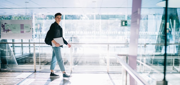 Side view of handsome young male in trendy jacket and denim carrying backpack and laptop while walk