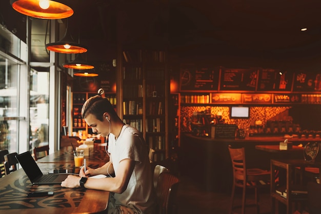 Side view of handsome young man in casual clothes with creative haircut sitting at window table in earphones with laptop and writing into notepad in bar