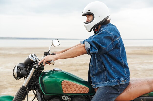 Side view of a handsome young man wearing casual outfit sitting on a motocycle at the beach, wearing a helmet