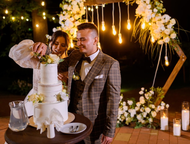 Side view of happy bride and groom standing near decorated arch together cutting wedding cake during night ceremony