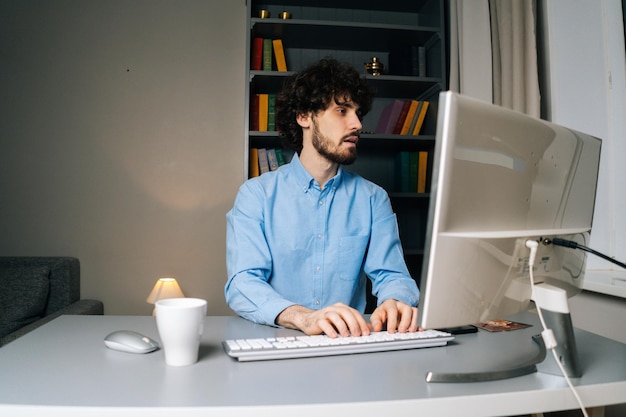 Side view of happy curly young man looking at camera and typing online message using computer