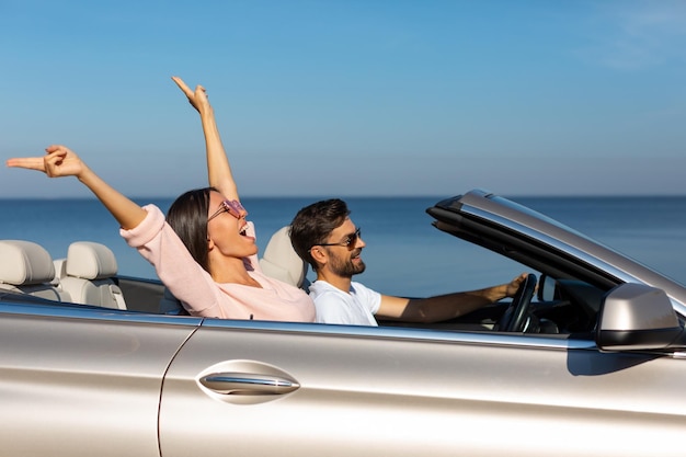 Side view of happy woman holding hands up sitting in cabriolet with boyfriend