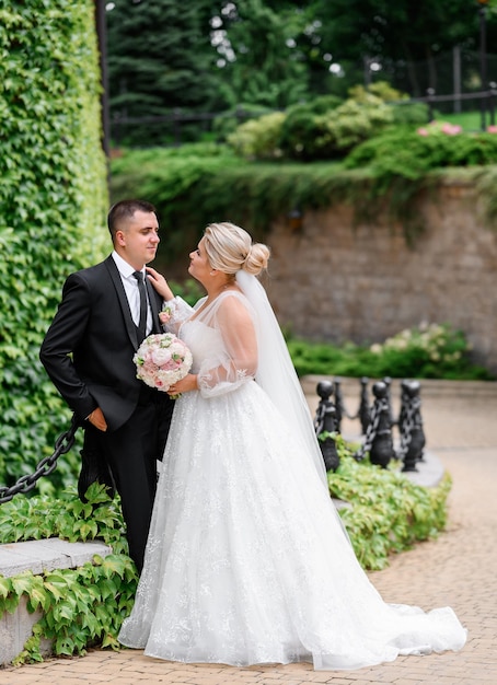 Side view of joyful man in wedding outfit keeping hands in pocket and leaning on decorated column smiling and looking to bride which standing opposite of him with bouquet of flowers