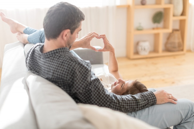 Side view of loving couple relaxing on couch and stacking hands in heart heart sign at home