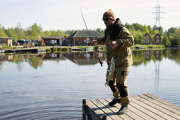 Side view of male fisher fishing with rod while standing on wooden quay near pond in countryside