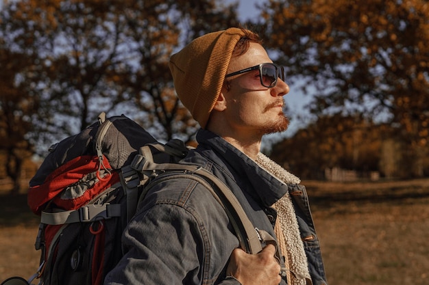 Photo side view of male traveler with hiking backpack in sunglasses and hipster wear standing on sunny day