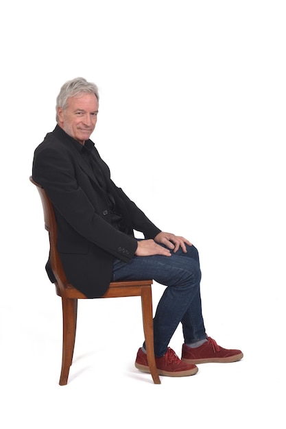 Side view of a man sitting on chair with sneakers jeans and blazer looking at camera on white background