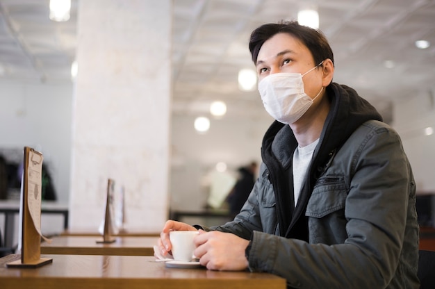 Side view of man with medical mask sitting at a table to have coffee