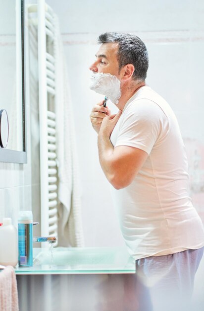 Side view of mature gray haired male shaving face with razor above sink while looking at himself in mirror