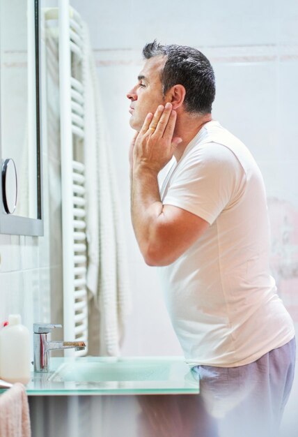 Side view of mature man wearing domestic clothes stroking face with cream during hygienic routine in bathroom while looking at himself in mirror