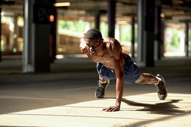 Side view of muscular black man in sportswear doing push ups during workout on street of modern downtown with urban background