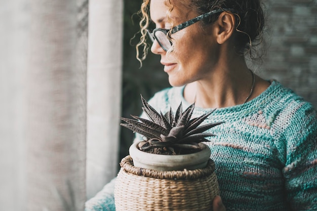 Side view of one pensive woman holding plant inside home and looking outside the window alone Female people wearing eyewear and caring interior garden nature Serene people feeling emotion Sadness