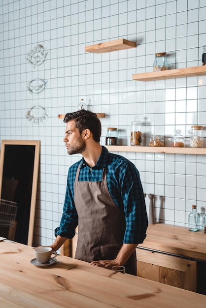 Side view of pensive barista standing at counter with cup of coffee in cafe