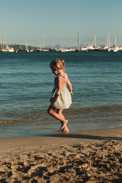 Side view of playful kid in blue dress walking on sandy beach near waving sea with yachts in dusk