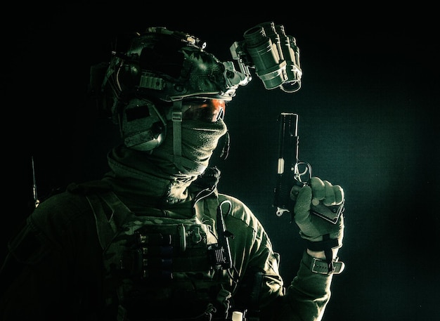 Side view portrait of army soldier modern combatant special forces fighter in helmet nightvision device radio headset hiding identity behind mask armed service pistol low key studio shoot