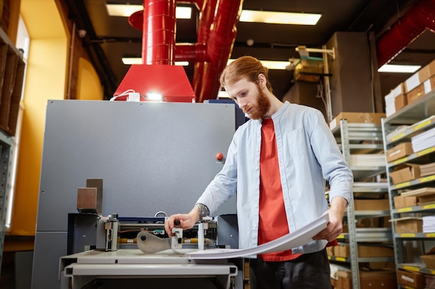 Side view portrait of bearded man putting paper in industrial printing machine at workshop copy spac