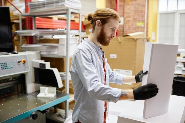 Side view portrait of bearded young man holding stack of paper while working in print shop copy spac