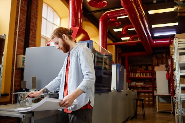 Side view portrait of bearded young man operating industrial printing machine at workshop copy space