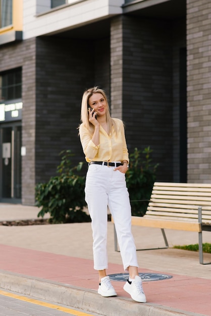 Side view portrait of a beautiful blonde woman holding a phone and laughing while talking on the phone against the background of the city