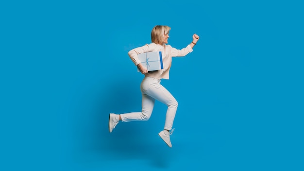 Side view portrait of a beautiful young woman running with a blue gift box against a blue wall. Fast delivery concept.