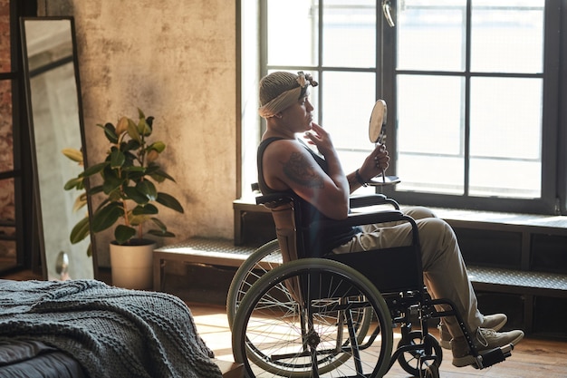 Side view portrait of black woman in wheelchair putting on makeup while looking at mirror in home interior